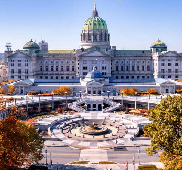 A gray state capitol building with a green roof with a water fountain out front