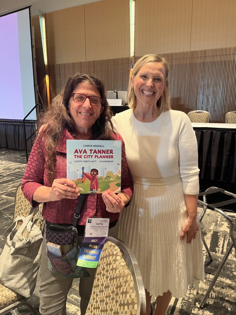 A brunette woman in a burgundy jacket and a blonde woman in a white dress hold a book called Ava Tanner The City Planner