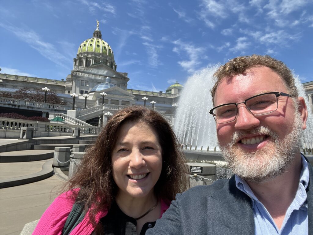 A brunette woman in a pink blazer and a man with glasses stand with the Pennsylvania Capitol behind them.